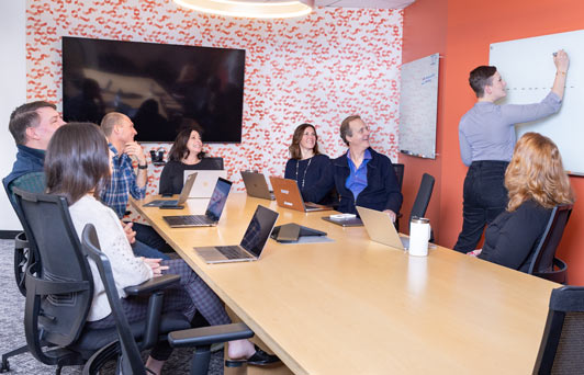 A group is sitting around a conference with room with one person at the white board, laying out a leadership development coaching strategy for his org.