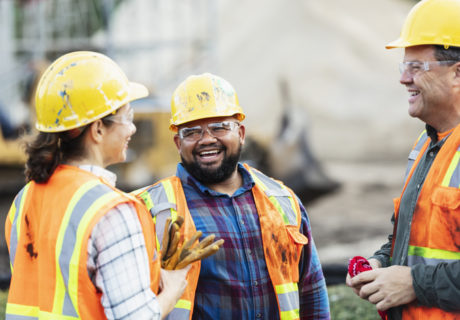 A group of three multi-ethnic workers at a construction site wearing hard hats, safety glasses and reflective clothing, smiling and conversing. The main focus is on the mixed race African-American and Pacific Islander man in the middle. The other two construction workers, including the woman, are Hispanic.